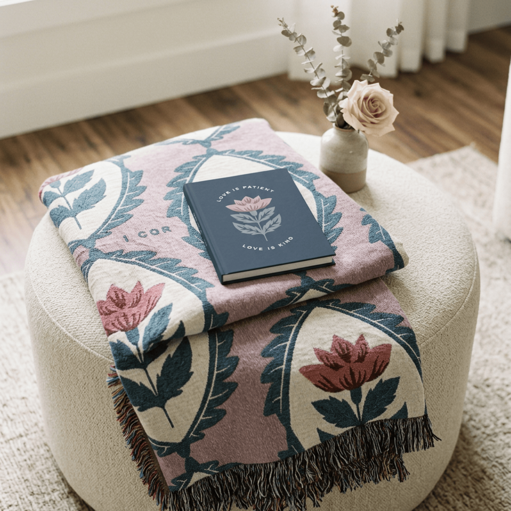 A patterned throw blanket with a dusty pink base, cream sections, and blue-green floral motifs is draped over a round beige ottoman. A navy book and a small vase with dried stems and a pale pink rose sit on top of the blanket in a softly styled room setting.