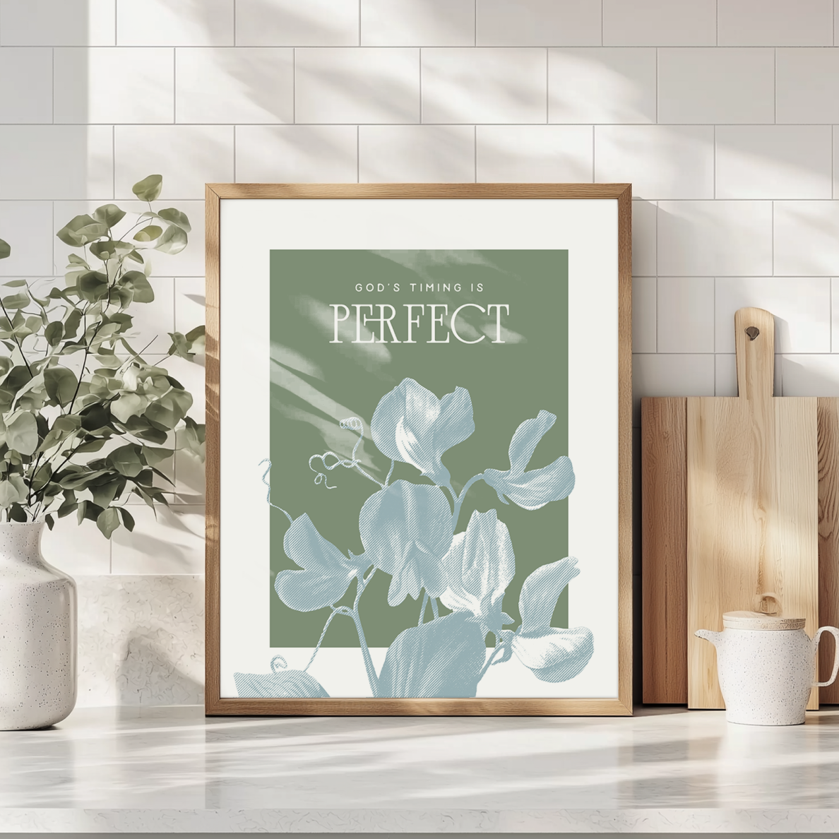 Framed artwork with 'God's Timing is Perfect' text on a green background, placed on a surface with a plant and books in the background.