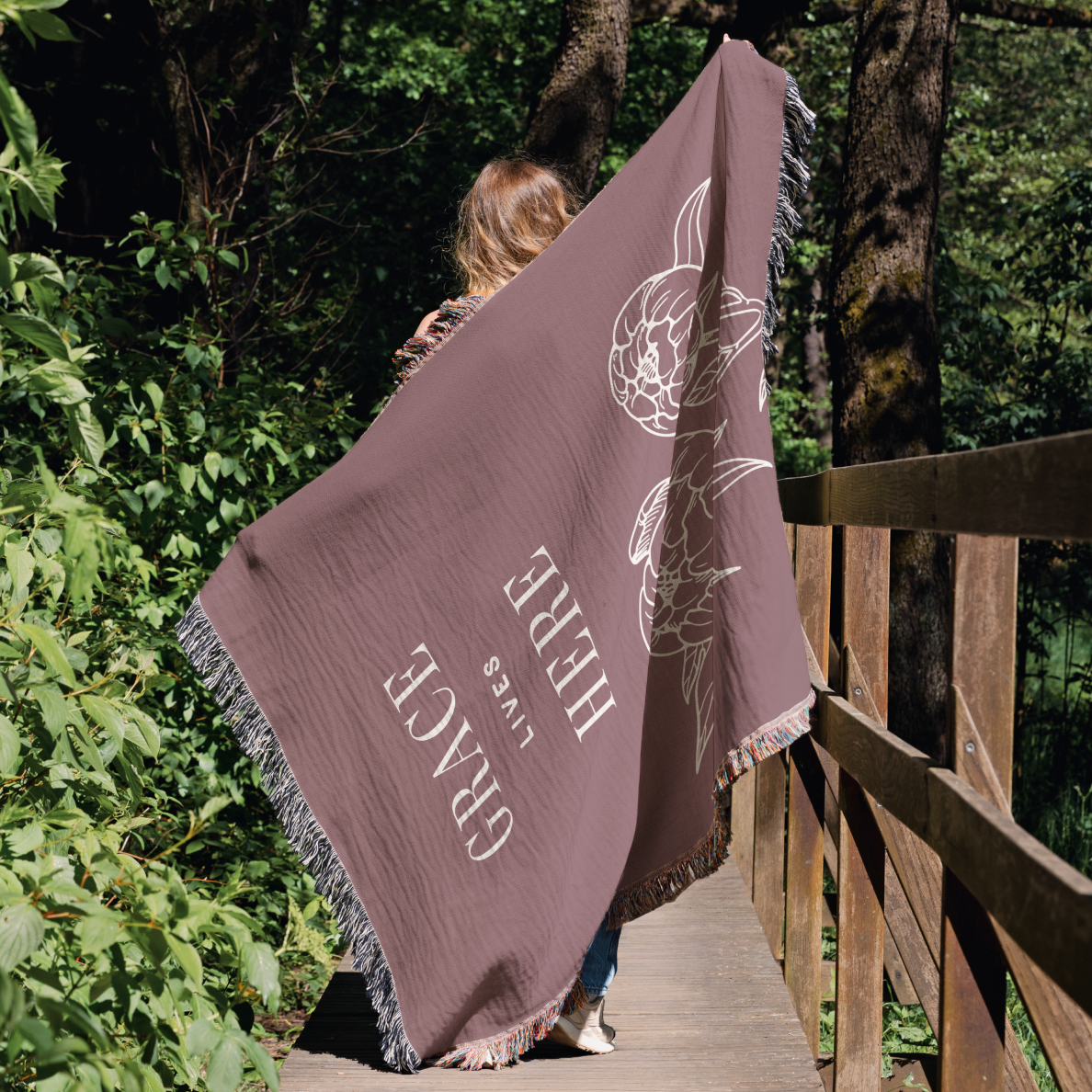 Person holding a large brown blanket with text and a design on a wooden bridge in a forest.