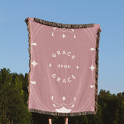 Pink blanket with 'Grace Upon Grace' text against a blue sky and green trees background
