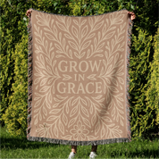 Neutral-toned woven blanket with the phrase "Grow in Grace" surrounded by elegant floral design; made from 100% cotton with fringe, perfect Christian gift or decor.