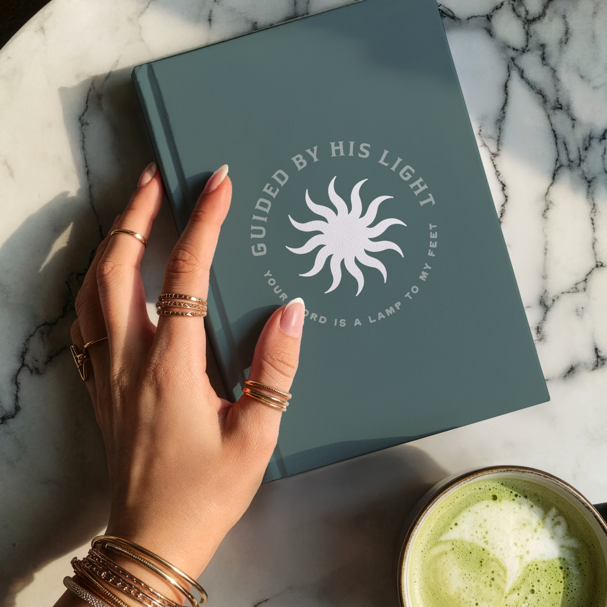 Hand holding a teal notebook with a sun design and text on a marble surface with a cup of green tea.