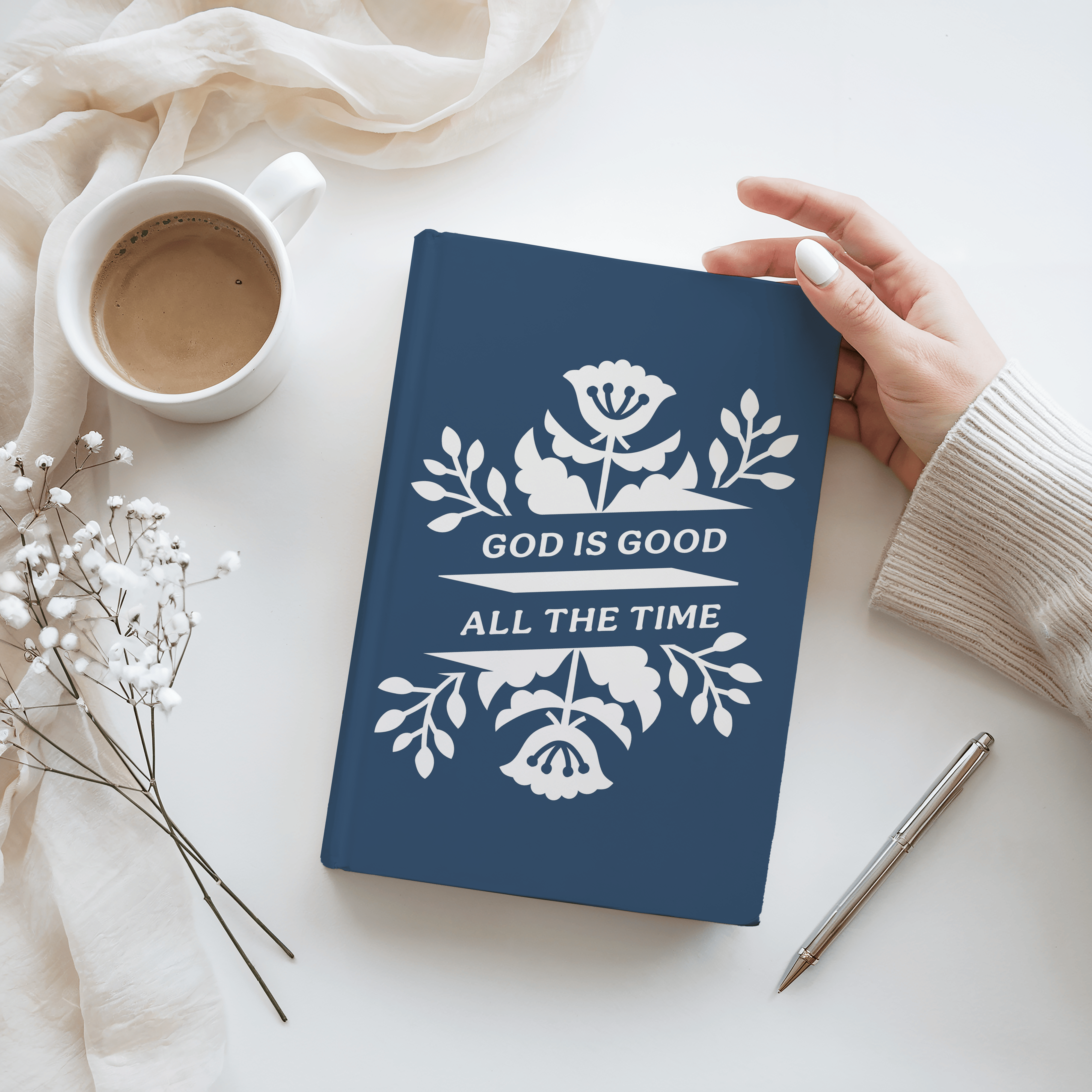 Blue notebook with 'God is Good All the Time' text held by a hand on a white surface with a cup and pen.