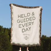 Blanket with text 'HELD & GUIDED EVERY DAY' against a blue sky and trees background