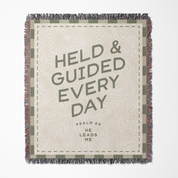 Blanket with text 'HELD & GUIDED EVERY DAY' and decorative border on a white background