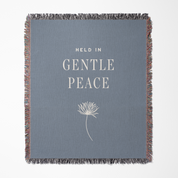 Gray blanket with 'Held in Gentle Peace' text and dandelion graphic on a white background