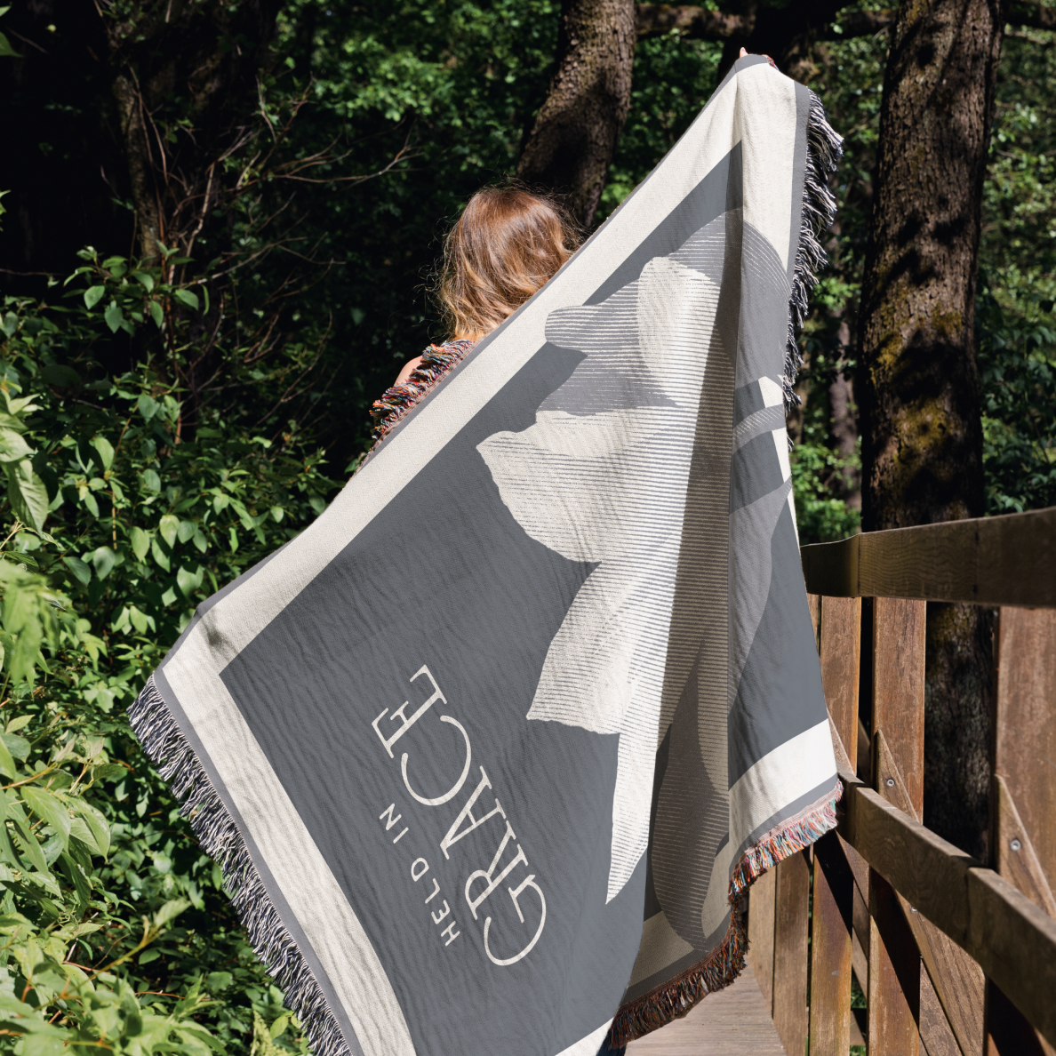 Person holding a large gray blanket with floral design and brand name in a forest setting