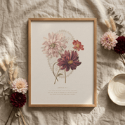 A framed botanical art print featuring pink and burgundy dahlia flowers on an off-white background, styled in a flat lay on a textured beige linen cloth with dried grasses, flowers, and a ceramic dish.