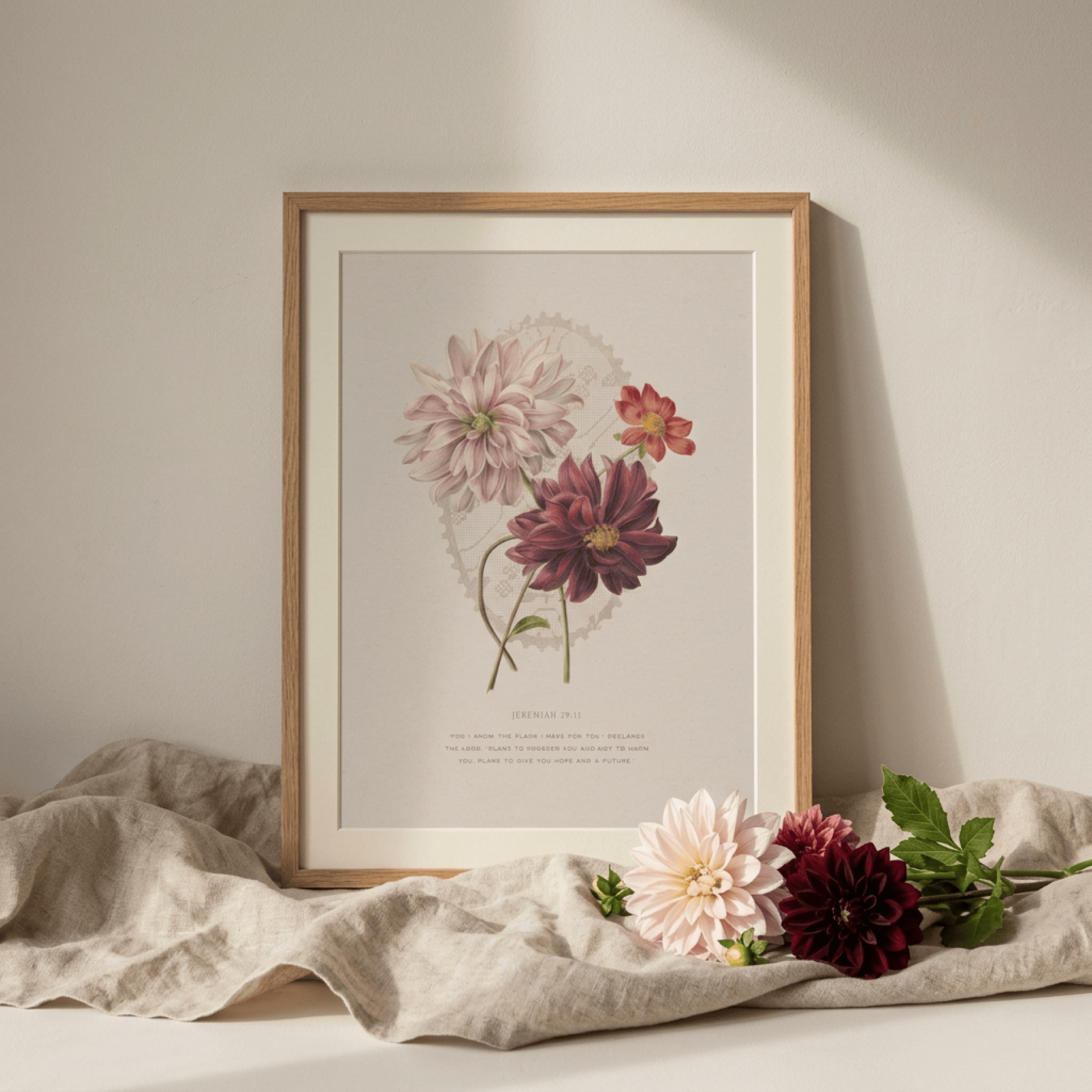 A framed botanical art print featuring vintage illustrations of dahlias, styled on a neutral beige surface with a draped linen cloth and fresh flowers in the foreground.