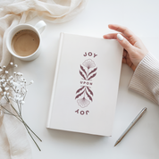 White notebook with 'JOY UPON YOU' design, cup of coffee, pen, and flowers on a white surface
