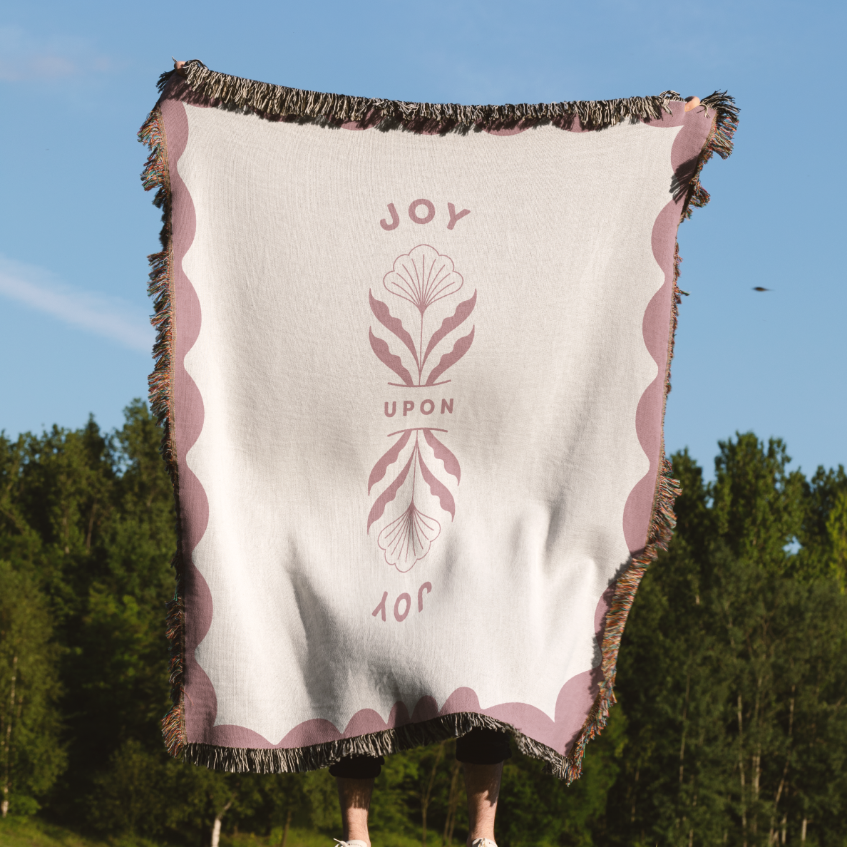 Blanket with 'JOY UPON' text and design, held up outdoors with trees and blue sky in the background