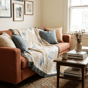 A cozy living room scene with an orange-brown upholstered sofa draped with a cream throw blanket featuring blue floral motifs and fringe. The blanket has visible text reading 'WORDS ARE THE HONEY'.