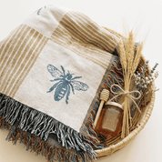 A woven throw blanket with a blue bee motif and multicolor fringe, styled in a wicker basket with dried wheat, a honey dipper, and a small honey jar on a light beige background.
