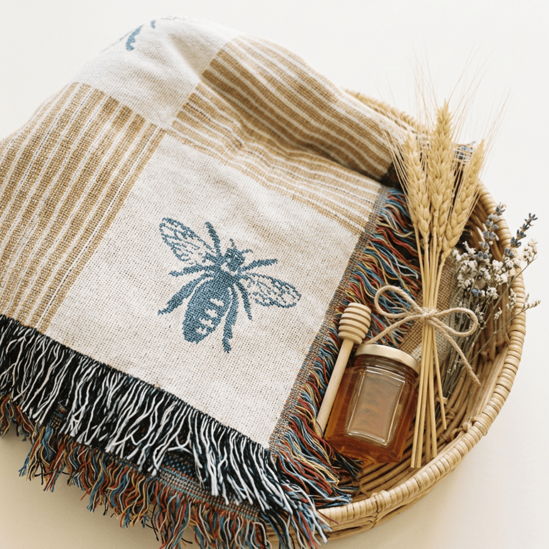 A woven throw blanket with a blue bee motif and multicolor fringe, styled in a wicker basket with dried wheat, a honey dipper, and a small honey jar on a light beige background.