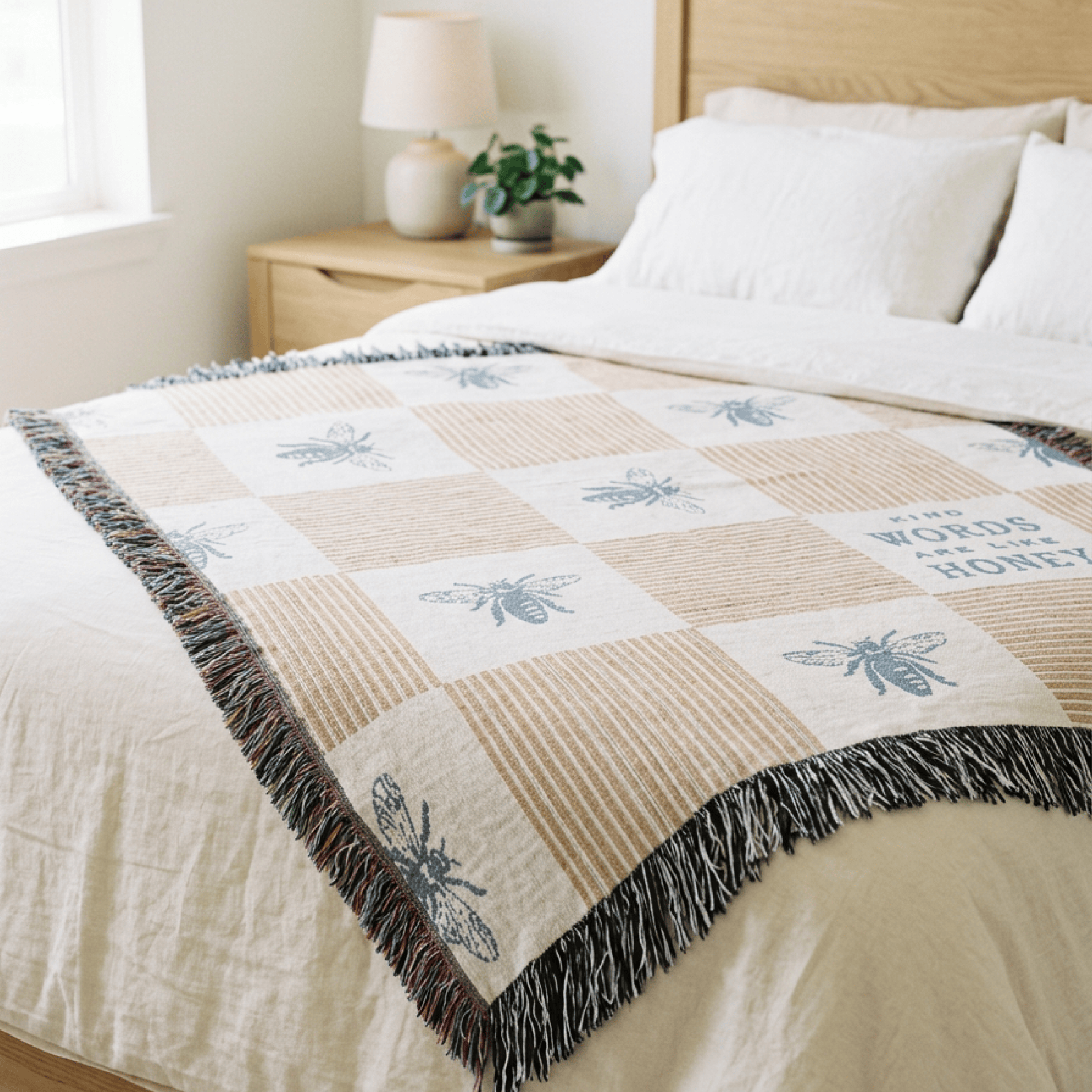 A woven throw blanket draped over a bed in a softly lit bedroom. The blanket has a cream and tan checkered pattern with blue-gray bee motifs, the phrase 'The words of honey' printed on one square, and dark multicolor fringe along the edges.