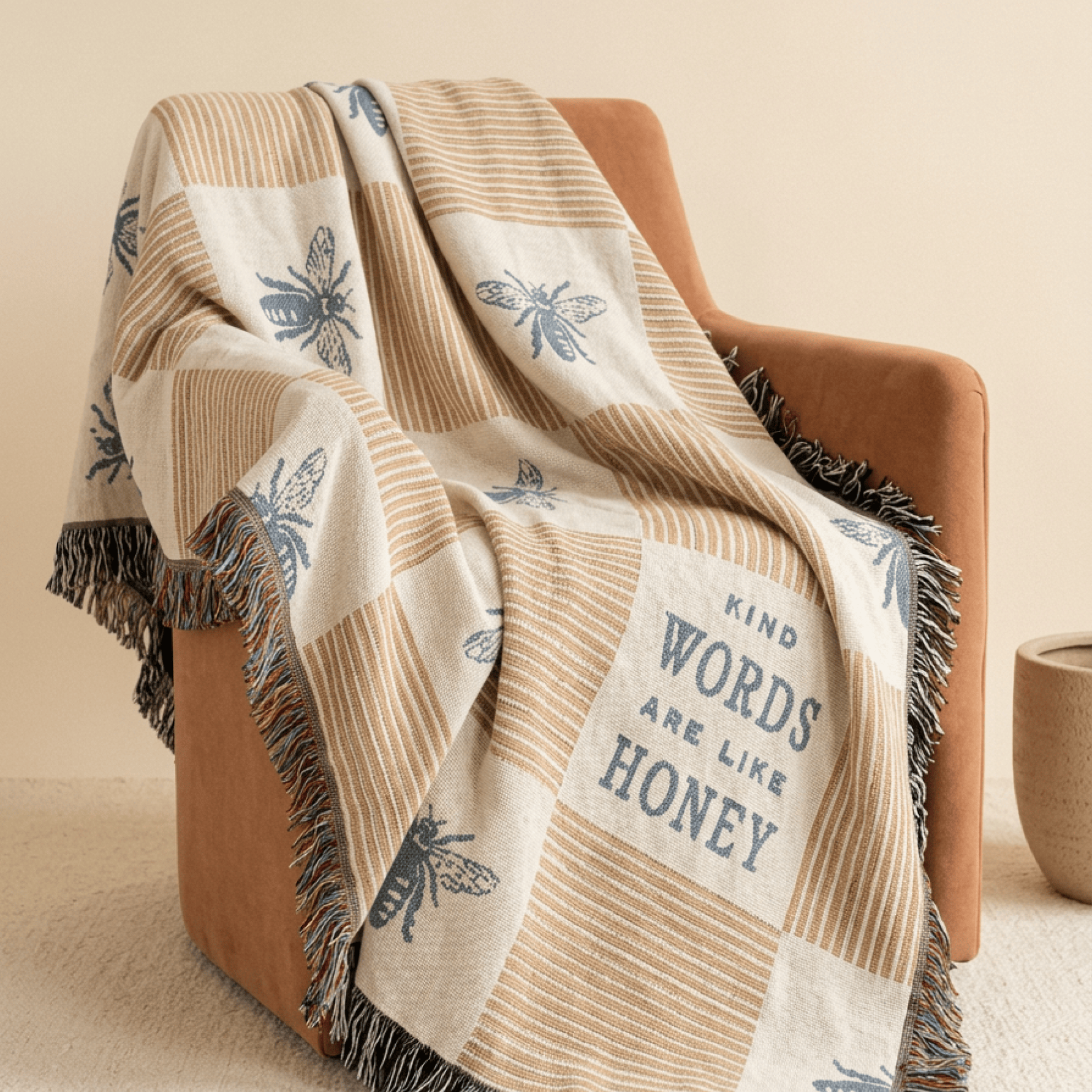 A woven throw blanket draped over a tan armchair in a softly lit room. The blanket has a cream and tan check pattern with blue bee motifs and the phrase “KIND WORDS ARE LIKE HONEY” printed on a cream panel.