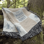 A woven throw blanket with fringe, draped over a mossy tree trunk outdoors. The blanket has a cream and tan striped pattern, blue bee motifs, and the text 'KIND WORDS ARE LIKE HONEY' printed in blue.