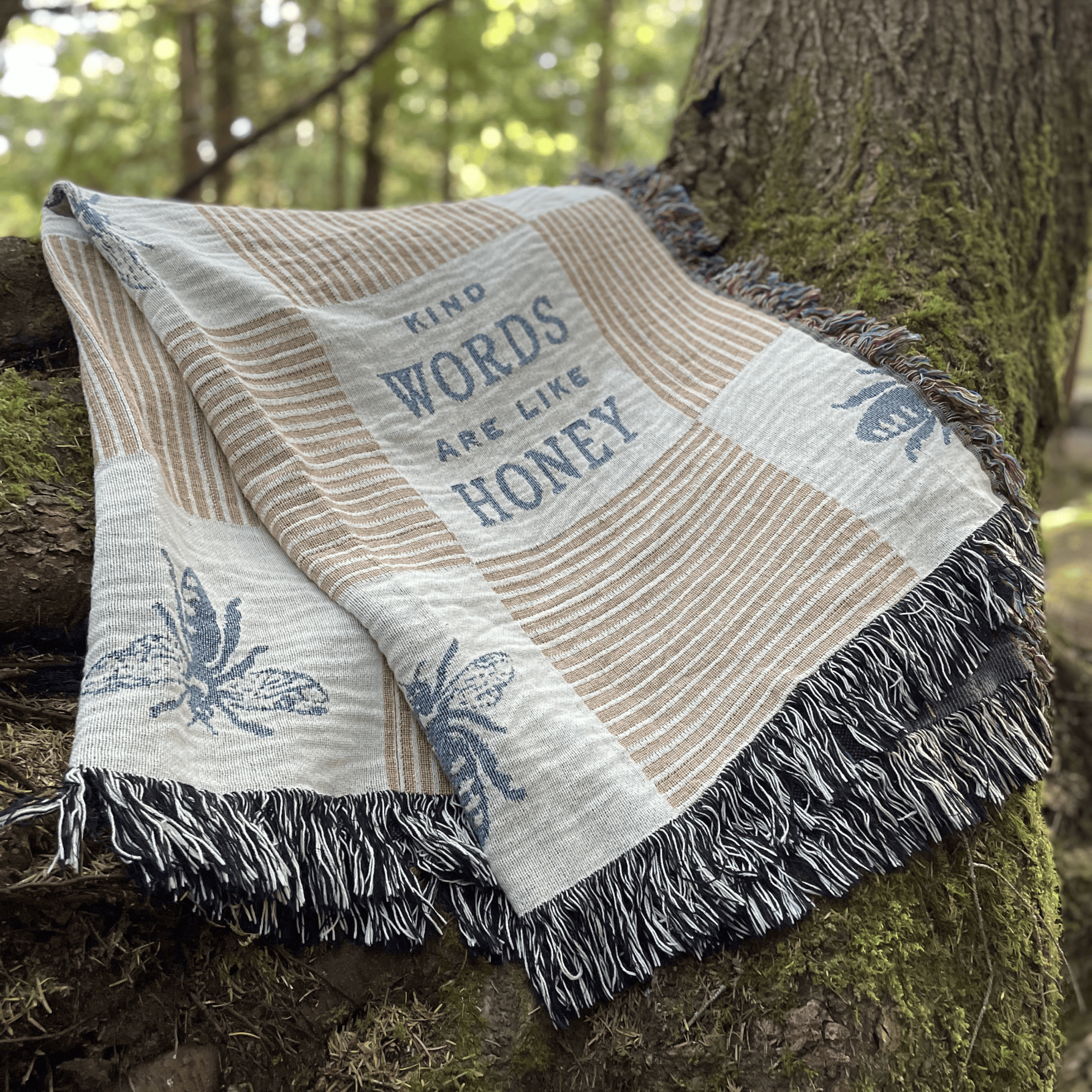 A woven throw blanket with fringe, draped over a mossy tree trunk outdoors. The blanket has a cream and tan striped pattern, blue bee motifs, and the text 'KIND WORDS ARE LIKE HONEY' printed in blue.
