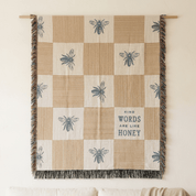 A woven wall tapestry with a checkered pattern of beige and cream squares, featuring blue-gray embroidered bees and stripes, fringe on the sides, and the text 'KIND WORDS ARE LIKE HONEY'.