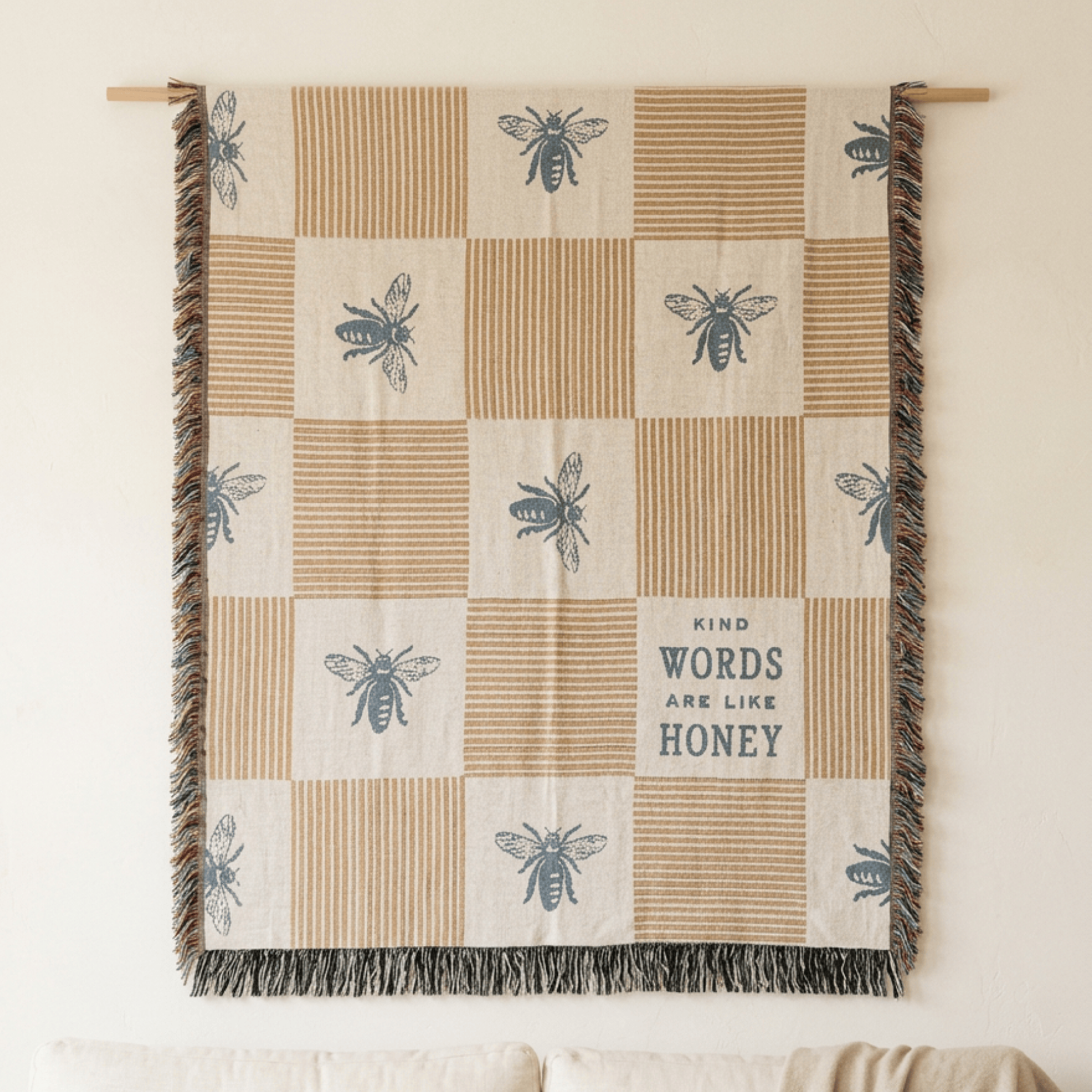 A woven wall tapestry with a checkered pattern of beige and cream squares, featuring blue-gray embroidered bees and stripes, fringe on the sides, and the text 'KIND WORDS ARE LIKE HONEY'.