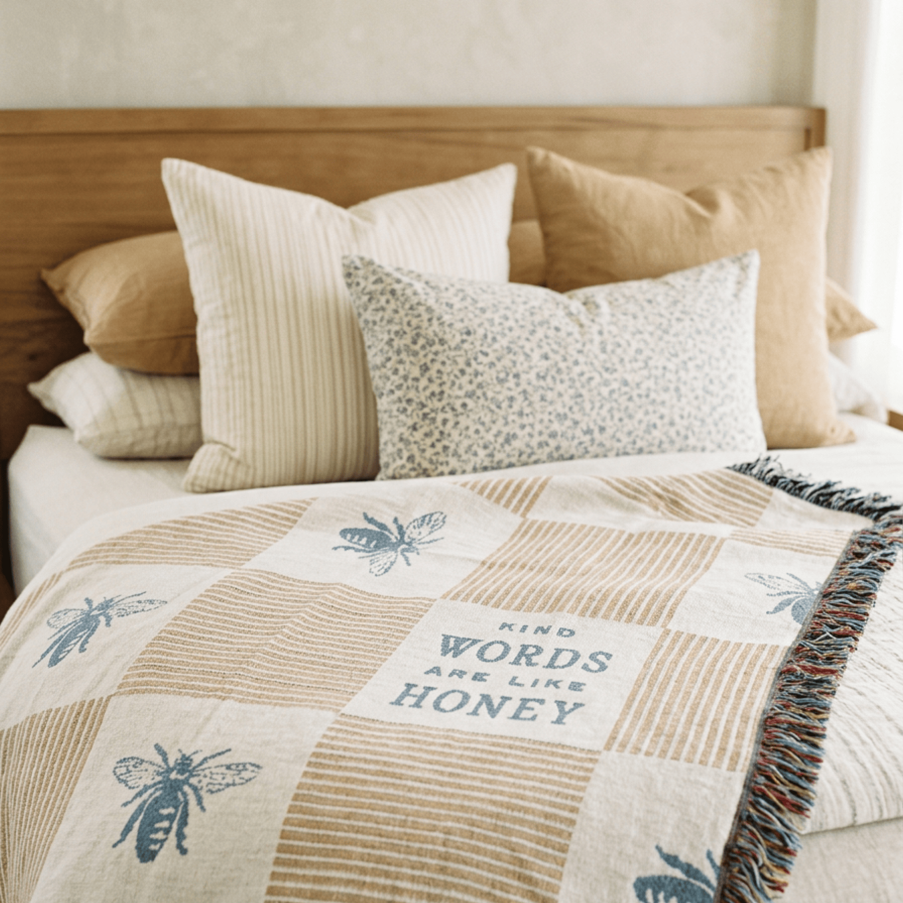 A cozy bed styled with a patchwork quilt featuring blue bee motifs and the phrase 'WORDS ARE LIKE HONEY' in a central square. The quilt is layered over neutral-toned bedding, including striped and floral accent pillows, set against a light wood headboard.