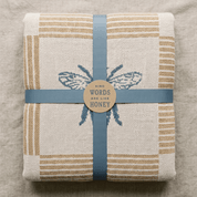 A folded woven textile, likely a towel or blanket, in a natural beige linen-like fabric with tan stripes and a blue embroidered bee motif. It is wrapped with a dusty blue ribbon and a round kraft paper label that reads 'KIND WORDS ARE LIKE HONEY'.