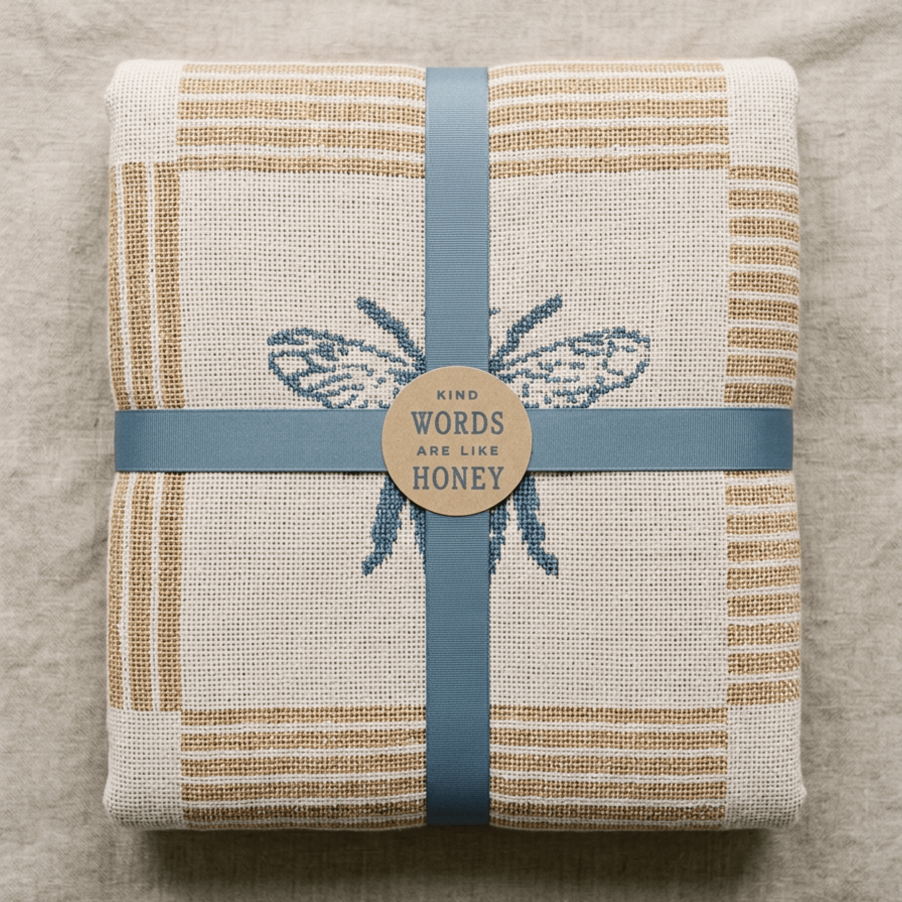 A folded woven textile, likely a towel or blanket, in a natural beige linen-like fabric with tan stripes and a blue embroidered bee motif. It is wrapped with a dusty blue ribbon and a round kraft paper label that reads 'KIND WORDS ARE LIKE HONEY'.