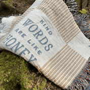 A woven throw blanket with the phrase 'KIND WORDS ARE LIKE HONEY' in blue lettering, laid outdoors on mossy ground. The blanket has a cream and tan striped pattern with blue text and multicolored fringe.