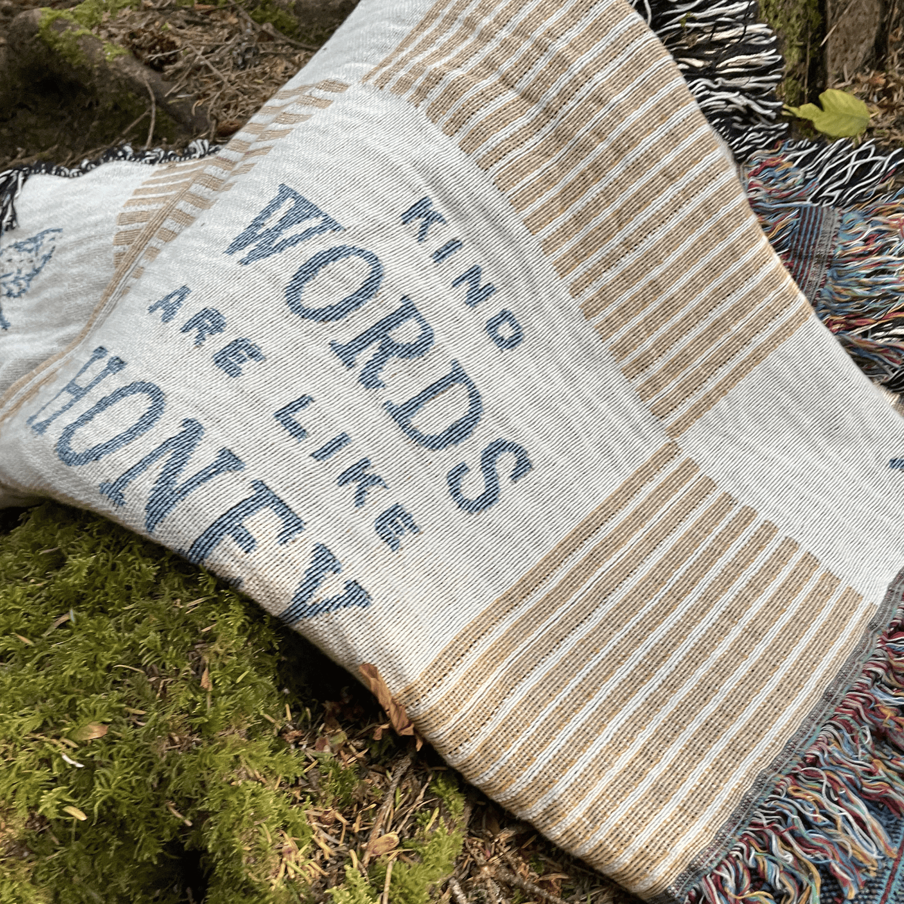 A woven throw blanket with the phrase 'KIND WORDS ARE LIKE HONEY' in blue lettering, laid outdoors on mossy ground. The blanket has a cream and tan striped pattern with blue text and multicolored fringe.