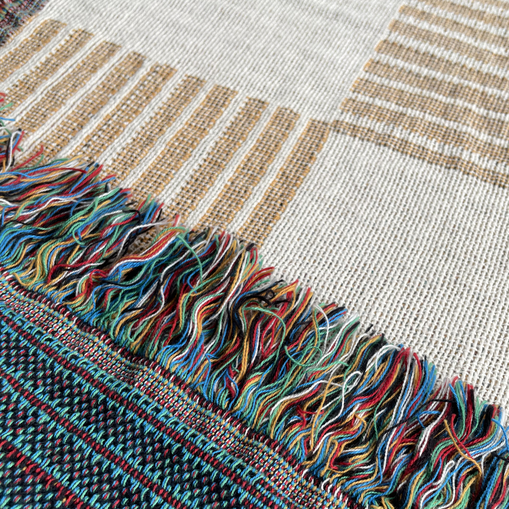 Close-up of a woven textile with a multicolored fringed edge, showing cream, tan, and dark navy striped sections with colorful thread tassels.