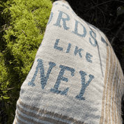 A close-up of a woven decorative pillow or cushion cover made from a natural beige burlap sack, featuring blue printed text that reads 'WORDS LIKE MONEY' and a rustic striped border. The pillow is photographed outdoors next to bright green moss.