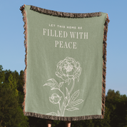 Green throw blanket with floral design and text 'Let this home be filled with peace' against a blue sky and tree background.
