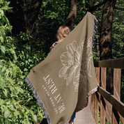 Person holding a brown blanket with floral design and text in a forest setting