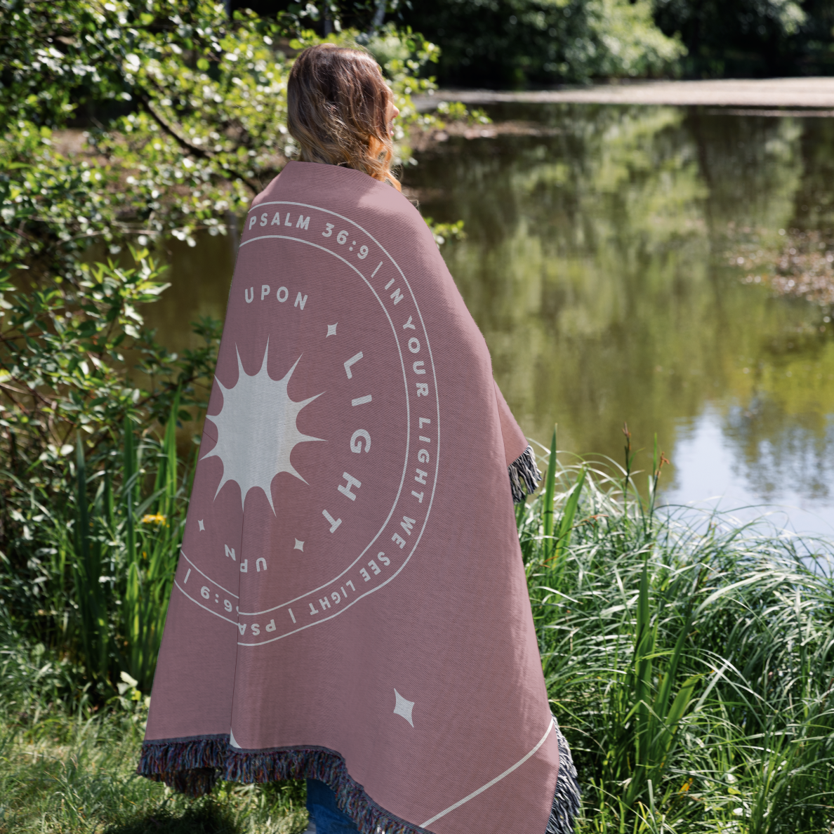Person wrapped in a pink blanket with text and sun design by a pond