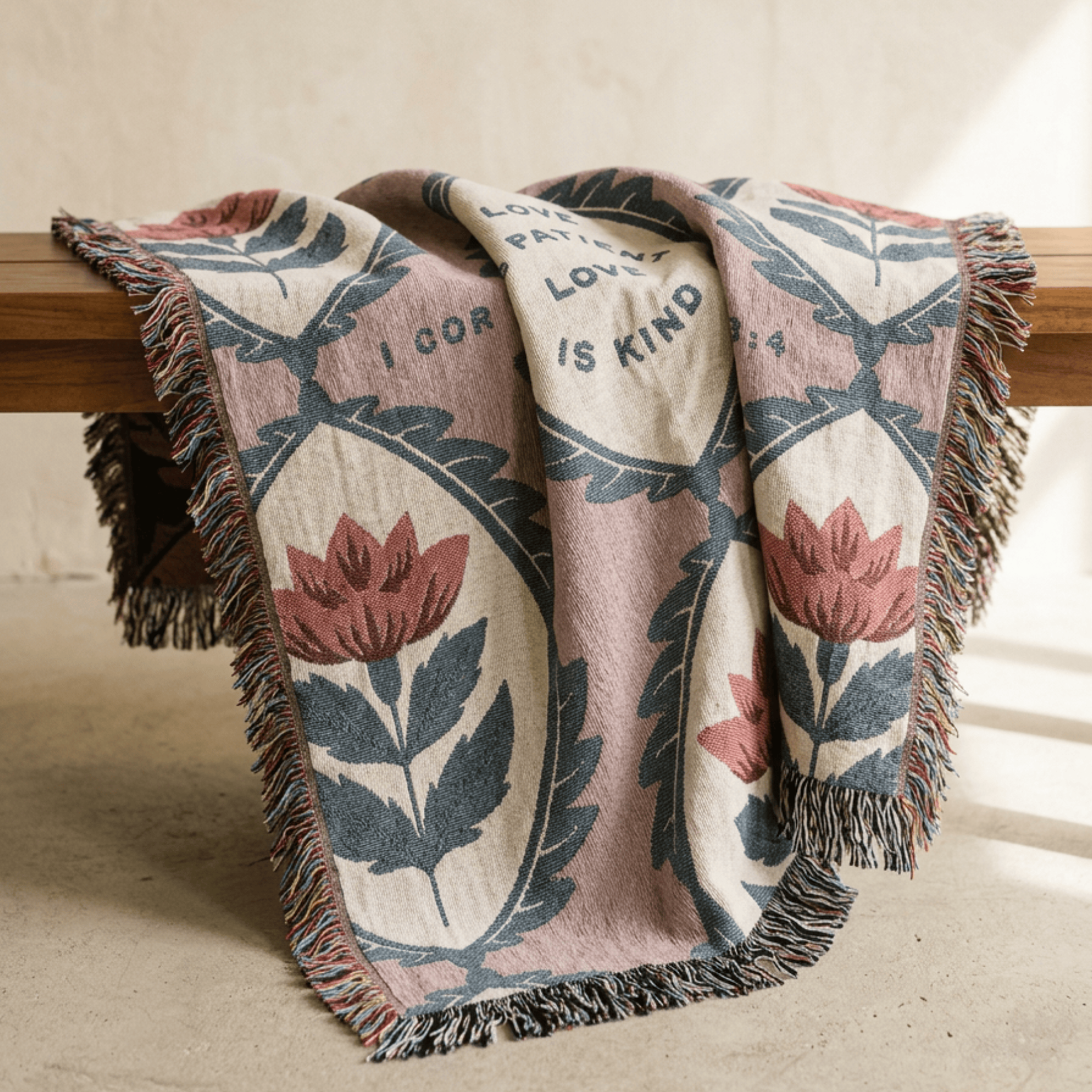 A woven throw blanket with fringe, draped over a wooden bench in a warm indoor setting. The blanket features a dusty pink background with cream panels, dark blue-gray leaf motifs, and muted red floral designs.