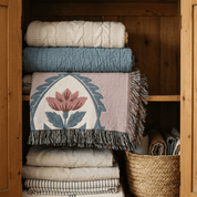 A woven throw blanket with a pink and blue lotus design and dark gray fringe, neatly folded on a wooden shelf alongside other home textiles.