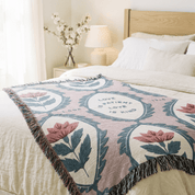 A woven throw blanket with a floral and shield pattern in dusty pink, cream, and slate blue, styled on a bed in a bright, modern bedroom.