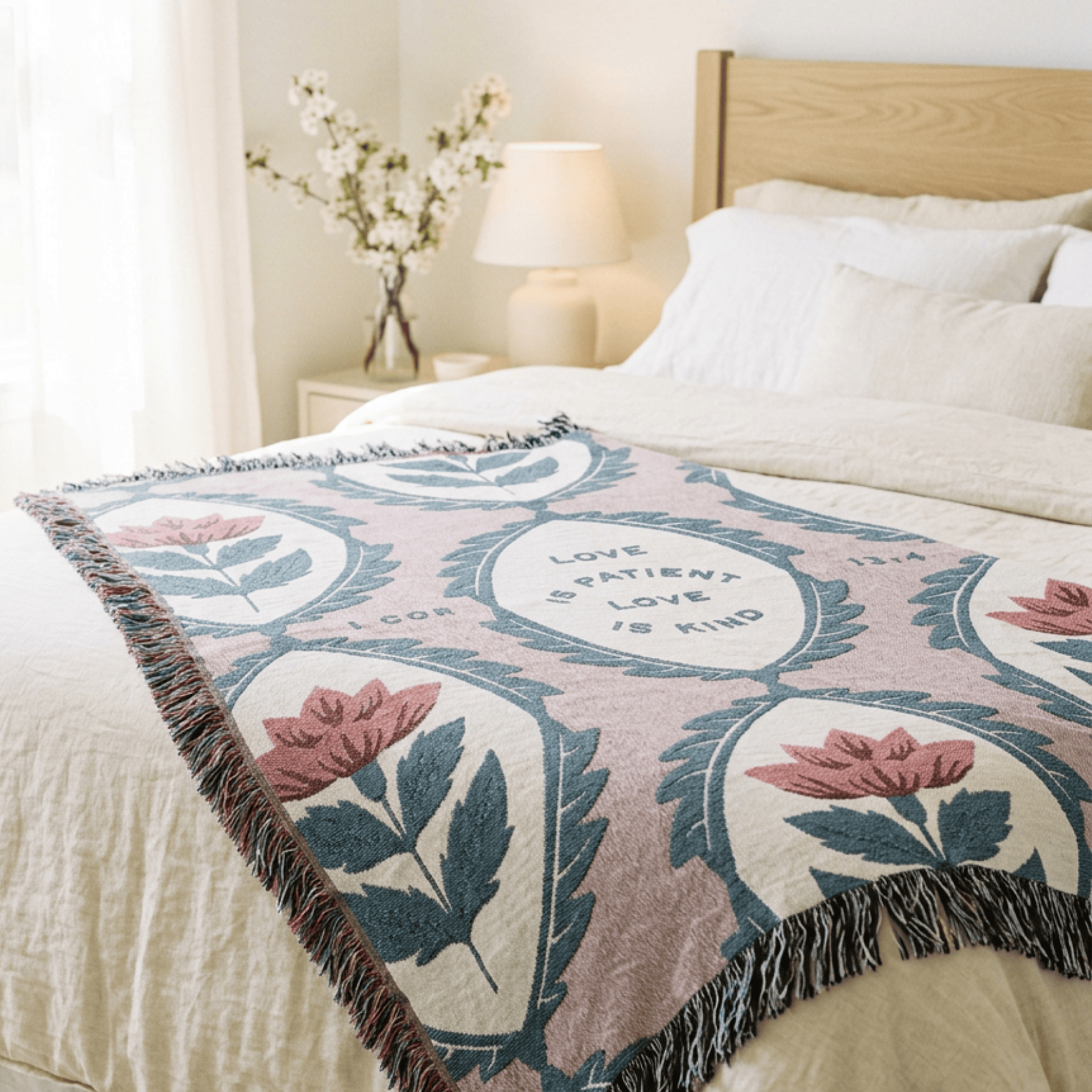 A woven throw blanket with a floral and shield pattern in dusty pink, cream, and slate blue, styled on a bed in a bright, modern bedroom.