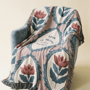 A woven throw blanket with a botanical pattern and fringe, draped over a blue-gray armchair against a warm beige background. The blanket features a dusty pink base with teal leaves and pink flowers, and a central white diamond containing the text 'LOVE IS PATRIOT'.