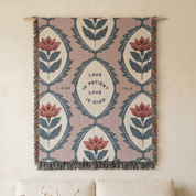 A woven wall tapestry with a botanical pattern and the words 'LOVE IS PATIENT LOVE IS KIND' from I Corinthians 16:14, hanging on a cream wall above a sofa.