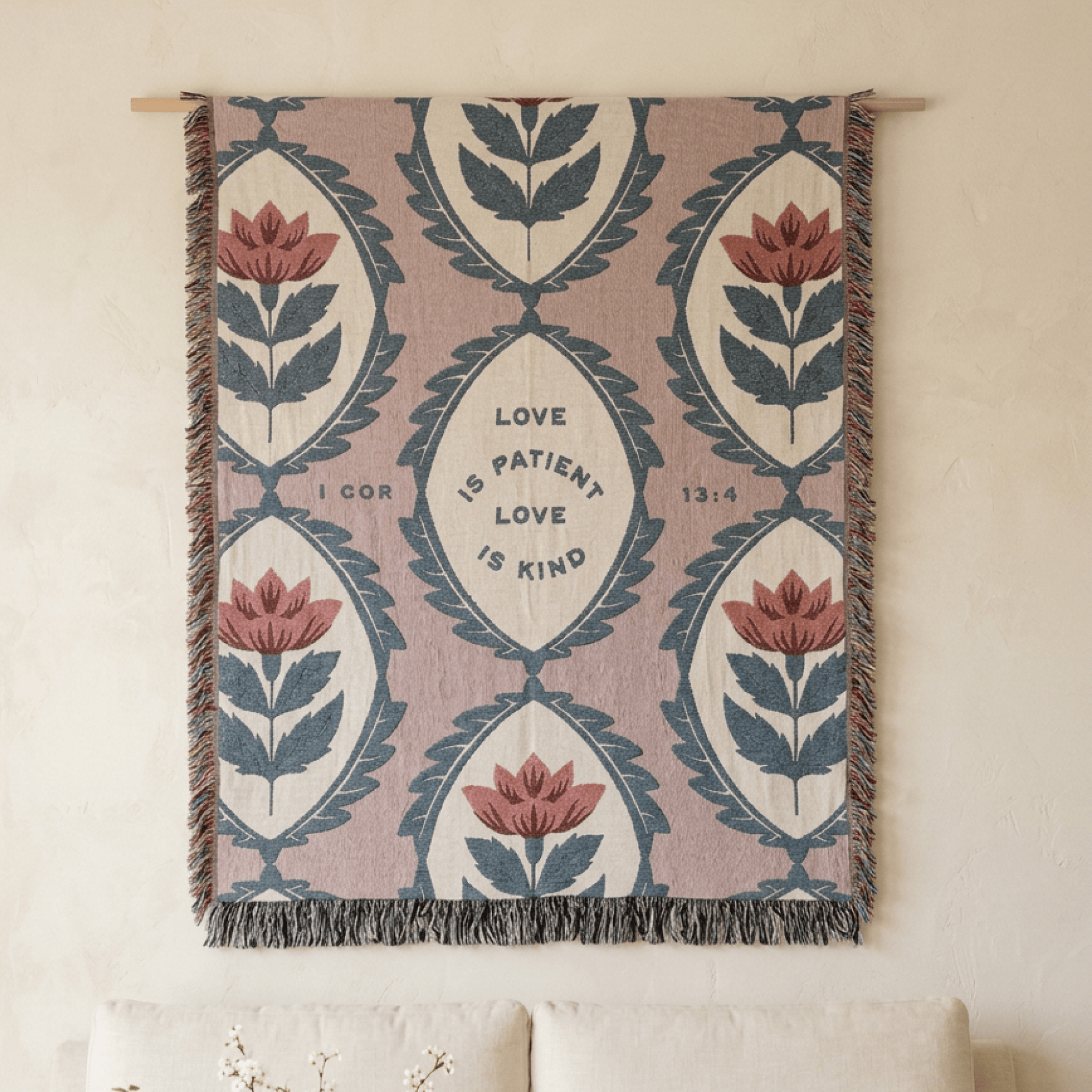 A woven wall tapestry with a botanical pattern and the words 'LOVE IS PATIENT LOVE IS KIND' from I Corinthians 16:14, hanging on a cream wall above a sofa.