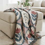 A woven throw blanket with a floral pattern and fringe, draped over a beige ottoman in a bright living room setting.