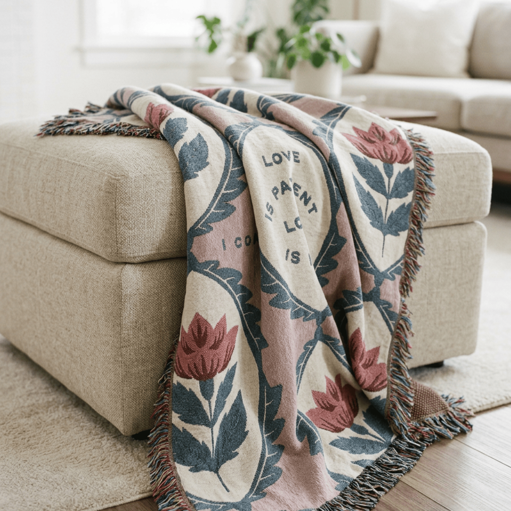 A woven throw blanket with a floral pattern and fringe, draped over a beige ottoman in a bright living room setting.