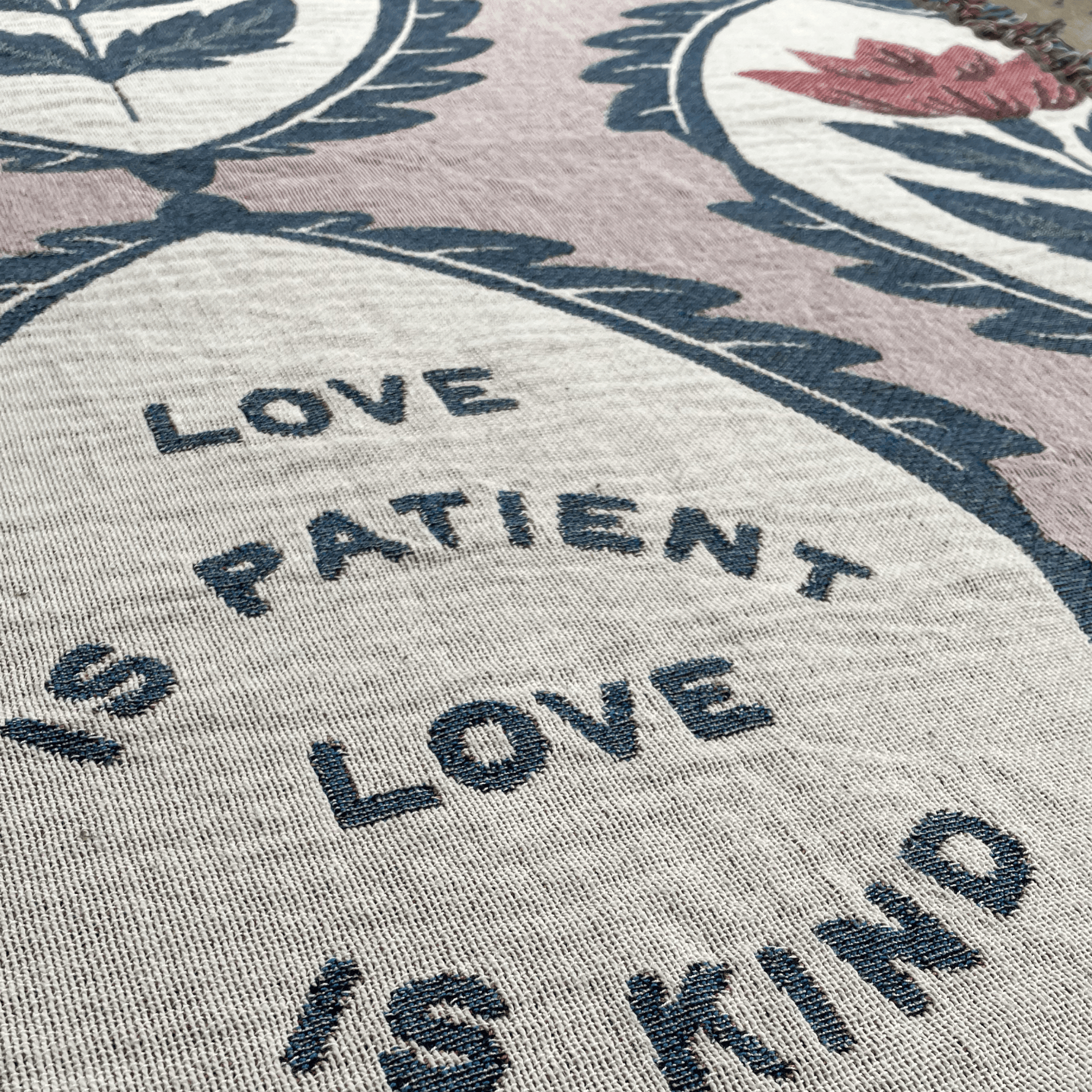 Close-up of a woven textile with the phrase “LOVE IS PATIENT LOVE IS KIND” in dark lettering on a light beige background, surrounded by floral and leaf motifs in muted pink, navy, and red.