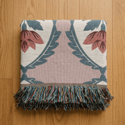 A square woven throw blanket with a floral and leaf pattern, featuring a dusty pink center, cream background, and blue-green accents, finished with a multicolor fringe and laid flat on a wooden floor.
