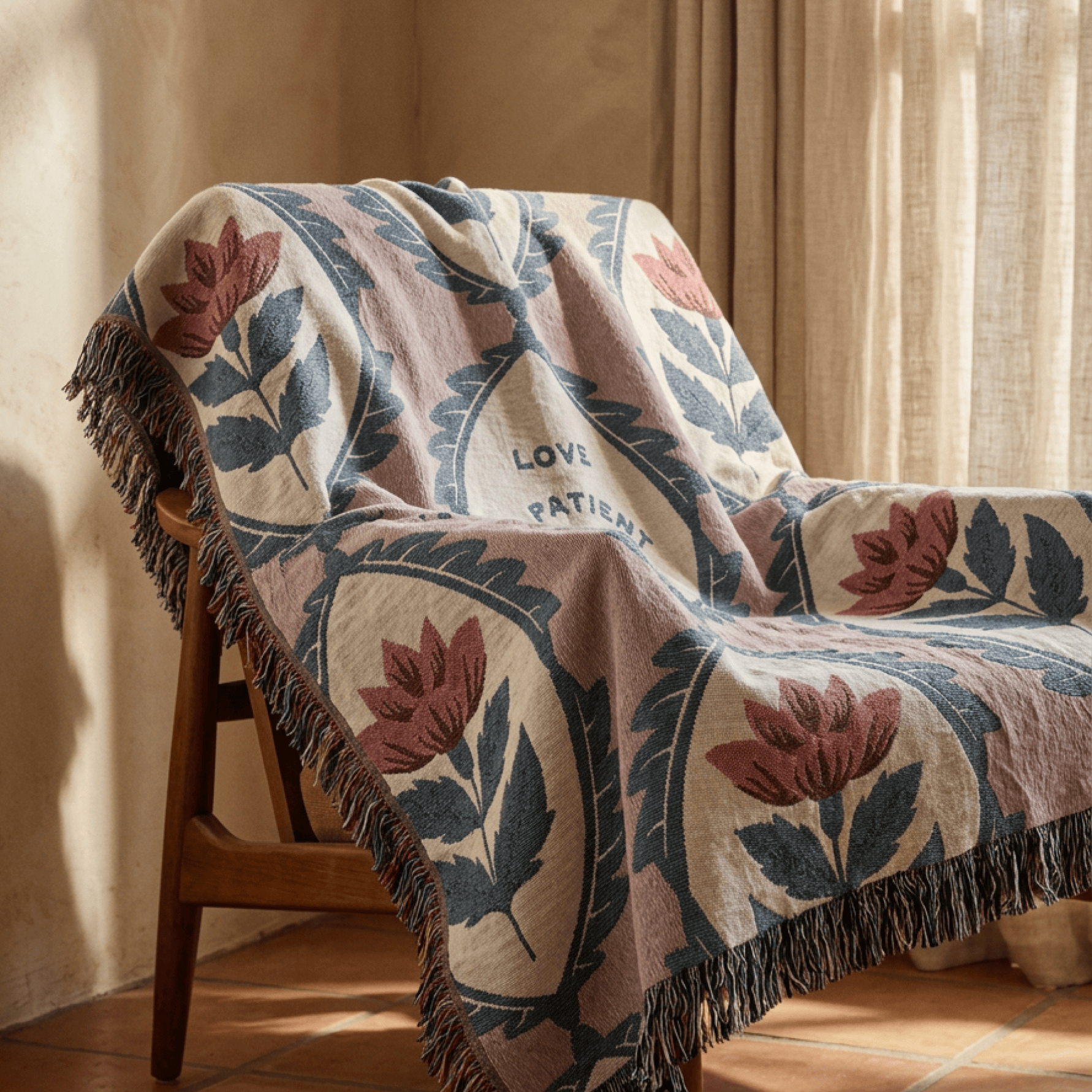 A patterned throw blanket draped over a wooden chair in a warm, sunlit room. The blanket features a cream base with a botanical design in dusty pink and slate blue, finished with dark fringe.