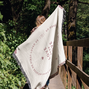 Person holding a white blanket with text and heart design in a forest setting