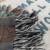 Close-up of a woven textile with thick black and white fringe, a multicolor striped woven section, and a light beige base printed with blue text.