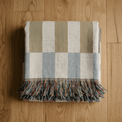 A folded woven throw blanket with a large check pattern in beige, light blue, and off-white, finished with a multicolor fringe, displayed on a light brown wooden floor.