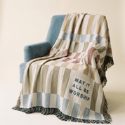 A multicolor striped woven throw blanket with fringe is draped over a blue armchair against a warm cream background. The blanket features a patchwork design in beige, blue, pink, and brown tones, with the text 'MAY IT ALL BE WORSHIP' printed on one section.